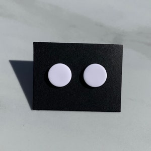 May include: Two white, circular stud earrings displayed on a black card. The earrings are a solid, light color and have a simple, minimalist design. The background is a light gray surface.