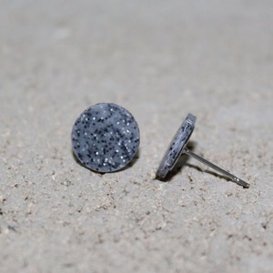 May include: A pair of gray stud earrings with a speckled design. The earrings are round and feature a silver post. One earring is upright, and the other is lying on its side.