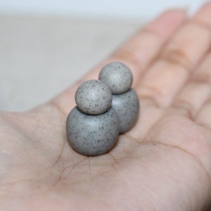 May include: Three small, speckled gray spheres stacked on top of each other, creating a unique shape. The spheres are resting in the palm of a hand, showcasing their size and texture. The gray color is consistent across all spheres.