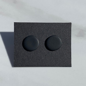 May include: A pair of matte black stud earrings. The earrings are circular and displayed on a black square card. The background is a light gray surface. These minimalist earrings are a simple accessory.