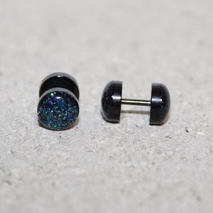 May include: A pair of circular stud earrings featuring a black metal frame and a dark, glittery surface with flecks of blue and green. One earring is shown from the front, while the other is positioned sideways to reveal the thin metal post.