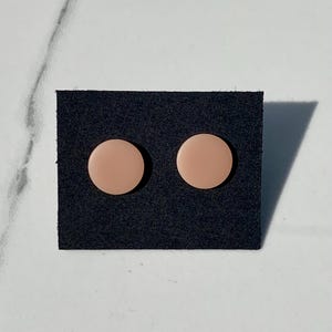 May include: A pair of blush-colored, circular stud earrings displayed on a black card. The earrings have a smooth, matte finish and are evenly spaced. The background is a white and gray marble surface.