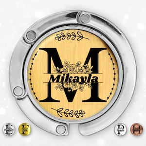 Puede incluir: Gancho plateado para bolso con un centro de madera que muestra el nombre "Mikayla" y la letra "M" en negro. El centro de madera tiene un diseño floral y está rodeado por un marco plateado. También se ven pequeños dijes.