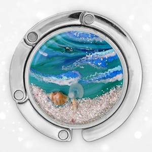 Puede incluir: Un gancho plateado para bolso con un diseño redondo con temática oceánica. El diseño presenta una escena de playa en miniatura con olas azules y verdes, una concha y arena. El gancho está diseñado para colgar un bolso de una mesa.
