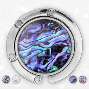 May include: A silver handbag hook featuring a round, iridescent abalone shell inlay. The hook has a circular design with a hinged opening and four small rivets. Additional abalone shell samples are displayed below the hook.