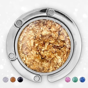 Puede incluir: Un gancho plateado para bolso con un diseño circular. El centro presenta un diseño de hoja de oro. El gancho tiene una forma curva con una pequeña abertura para colgar. Se muestran opciones de color adicionales debajo.