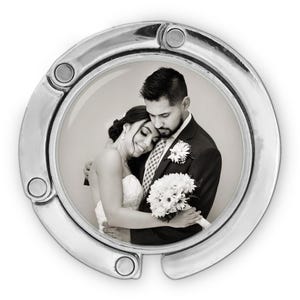 May include: A silver-colored purse hook with a black and white photograph of a couple embracing. The photograph is circular and shows a bride and groom on their wedding day. The hook has a curved design with a circular frame.