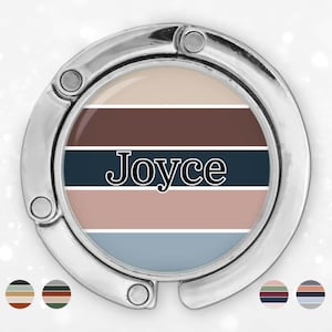 Puede incluir: Un gancho plateado para bolso con un diseño circular. El centro presenta rayas horizontales en tonos beige, marrón, azul marino, rosa y azul, con el nombre "Joyce" en negrita negra.