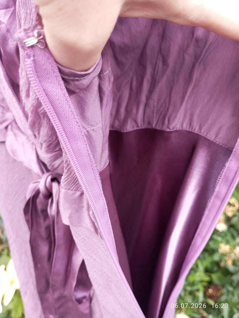 Puede incluir: Un vestido morado con cierre de cremallera lateral y un corchete de metal en la parte superior. La prenda incluye un cintur&oacute;n de tela a juego y un forro suave similar al sat&eacute;n visible en el interior, mostrando los detalles de confecci&oacute;n.