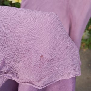 Puede incluir: Una tela de color morado claro con una textura ondulada y arrugada distintiva. El material presenta una peque&ntilde;a mancha circular de color marr&oacute;n rojizo oscuro situada cerca del centro de la secci&oacute;n visible. La tela parece ligera y semitransparente.