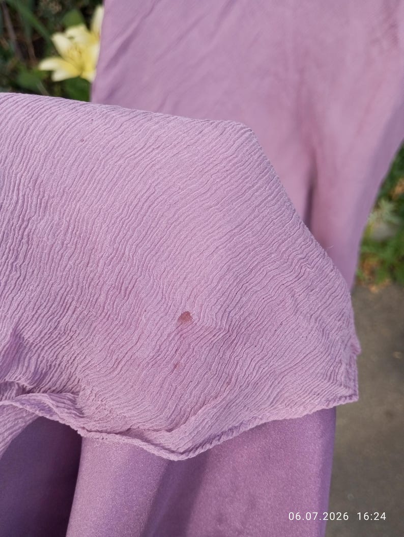 Puede incluir: Una tela de color morado claro con una textura ondulada y arrugada distintiva. El material presenta una peque&ntilde;a mancha circular de color marr&oacute;n rojizo oscuro situada cerca del centro de la secci&oacute;n visible. La tela parece ligera y semitransparente.