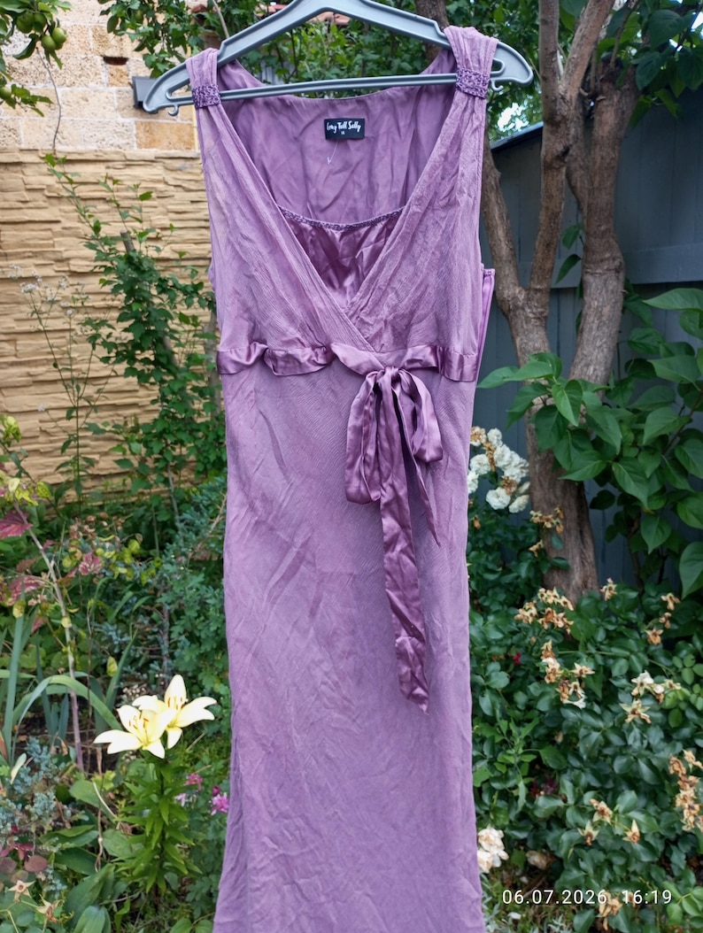 Puede incluir: Un vestido malva sin mangas con escote en V, cuerpo fruncido y un lazo de sat&eacute;n en la cintura. La prenda incluye una etiqueta que dice Long Tall Sally en el cuello interior y presenta detalles de tela texturizada en los tirantes.