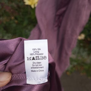 Puede incluir: Una prenda de color malva con una etiqueta de cuidado blanca que dice: 100% Silk, Lining: 100% Polyester, Dry clean, Do not iron embellishment, Made in China, junto con s&iacute;mbolos de cuidado est&aacute;ndar, incluido un icono de plancha tachado.