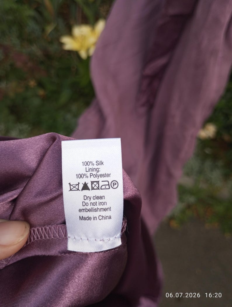 Puede incluir: Una prenda de color malva con una etiqueta de cuidado blanca que dice: 100% Silk, Lining: 100% Polyester, Dry clean, Do not iron embellishment, Made in China, junto con s&iacute;mbolos de cuidado est&aacute;ndar, incluido un icono de plancha tachado.