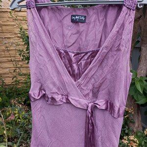 Puede incluir: Un vestido malva sin mangas con una capa de tela texturizada y arrugada y un forro de corpi&ntilde;o satinado. El vestido presenta un escote en V, una cinta de sat&eacute;n en la cintura y tirantes decorativos con cuentas. Una etiqueta dice Long Tall Sally.