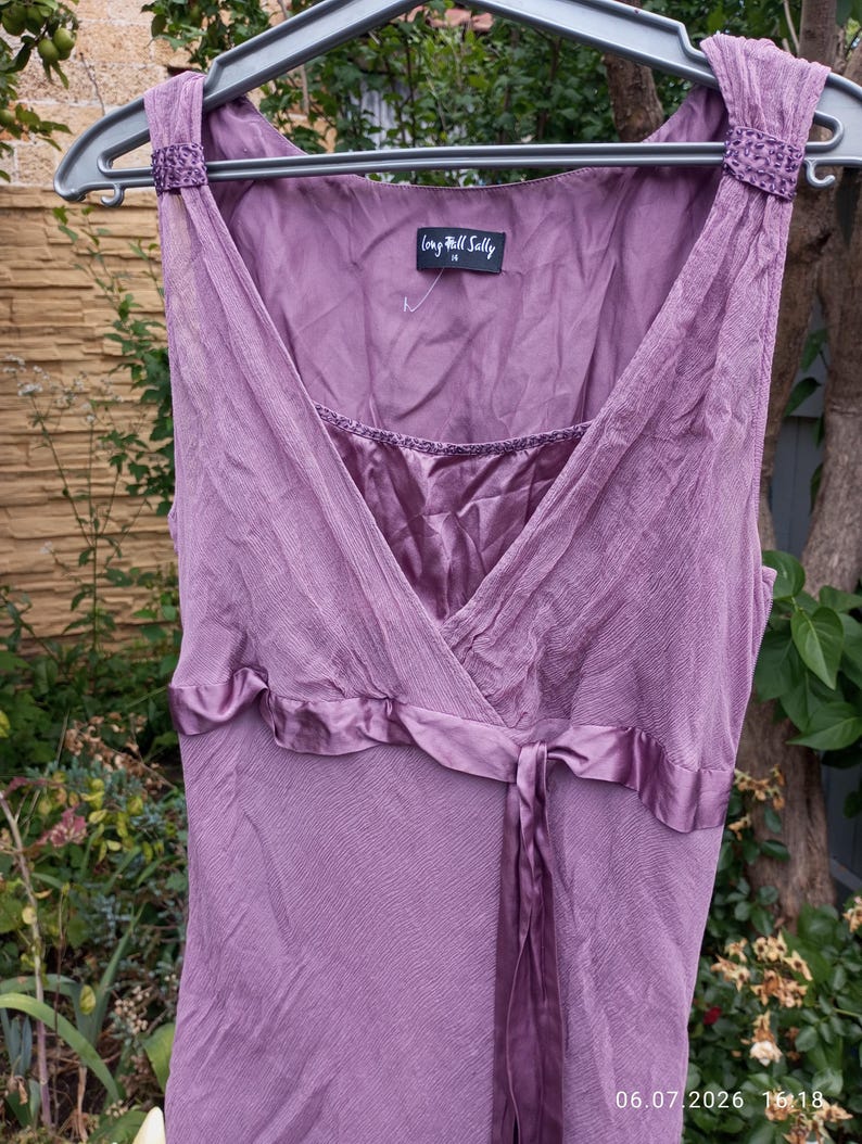 Puede incluir: Un vestido malva sin mangas con una capa de tela texturizada y arrugada y un forro de corpi&ntilde;o satinado. El vestido presenta un escote en V, una cinta de sat&eacute;n en la cintura y tirantes decorativos con cuentas. Una etiqueta dice Long Tall Sally.