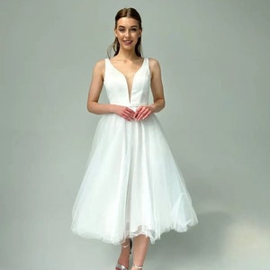 May include: A sleeveless white knee-length dress with a plunging neckline and a tulle skirt. The dress has a fitted bodice and a flowing skirt. The person is wearing silver high-heeled sandals.