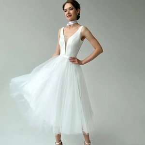 May include: A white knee-length dress with a V-neckline and a tulle skirt. The dress has a fitted bodice and a flowing skirt. The model is wearing white high-heeled sandals and a white choker. The dress is suitable for a wedding or special occasion.