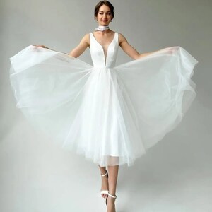 May include: A white knee-length dress with a plunging neckline and a flowing tulle skirt. The sleeveless dress is paired with white strappy heels. A white choker with a subtle pattern is worn around the neck.