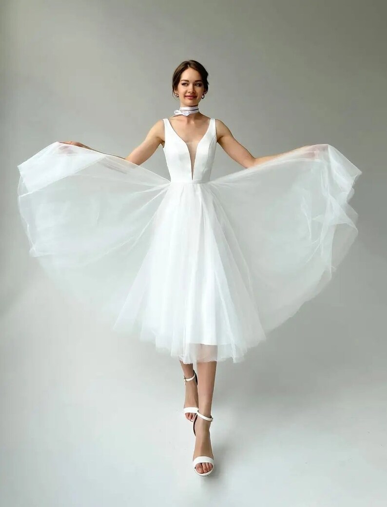 May include: A white knee-length dress with a plunging neckline and a flowing tulle skirt. The sleeveless dress is paired with white strappy heels. A white choker with a subtle pattern is worn around the neck.
