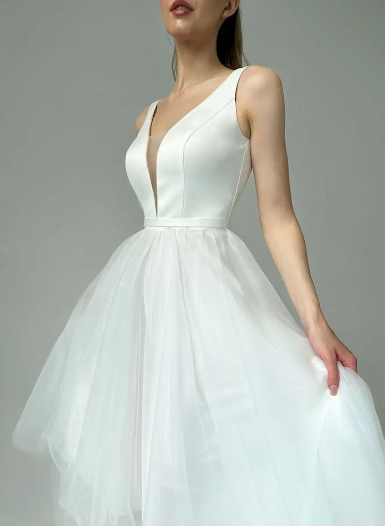 May include: A sleeveless white wedding dress with a deep V-neckline and a fitted bodice. The dress features a flowing tulle skirt and a thin belt at the waist. The fabric appears to be satin and tulle.