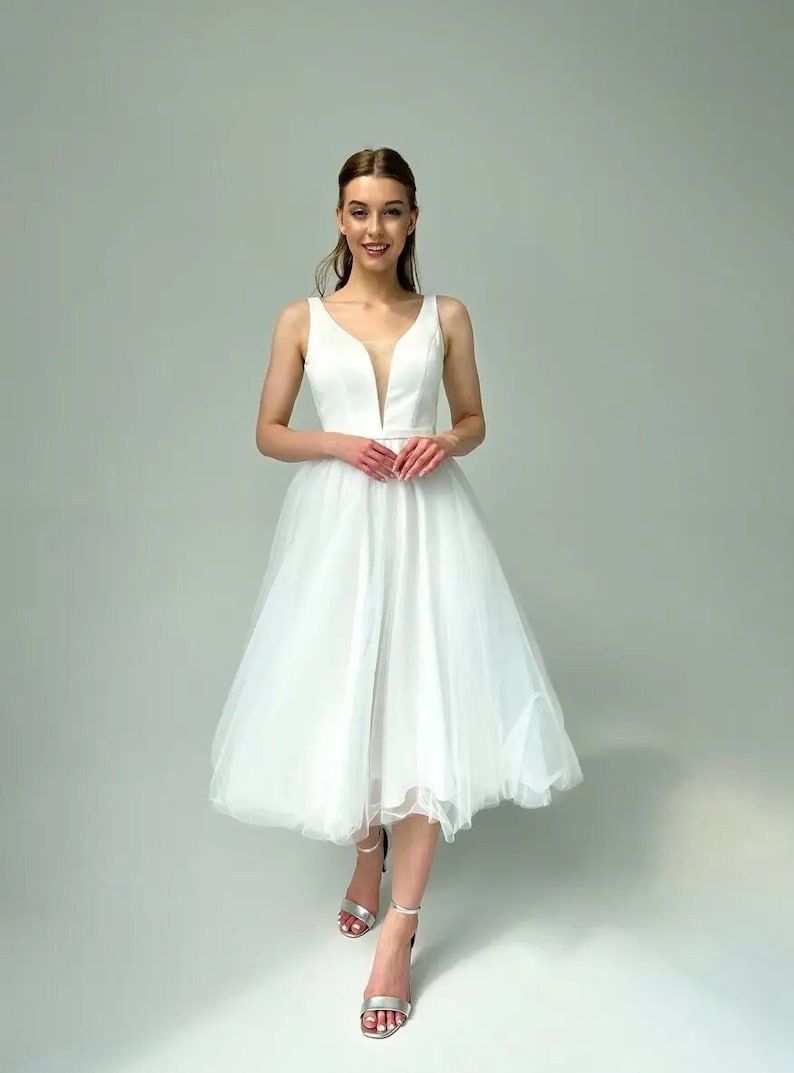 May include: A sleeveless white knee-length dress with a plunging neckline and a tulle skirt. The dress has a fitted bodice and a flowing skirt. The person is wearing silver high-heeled sandals.