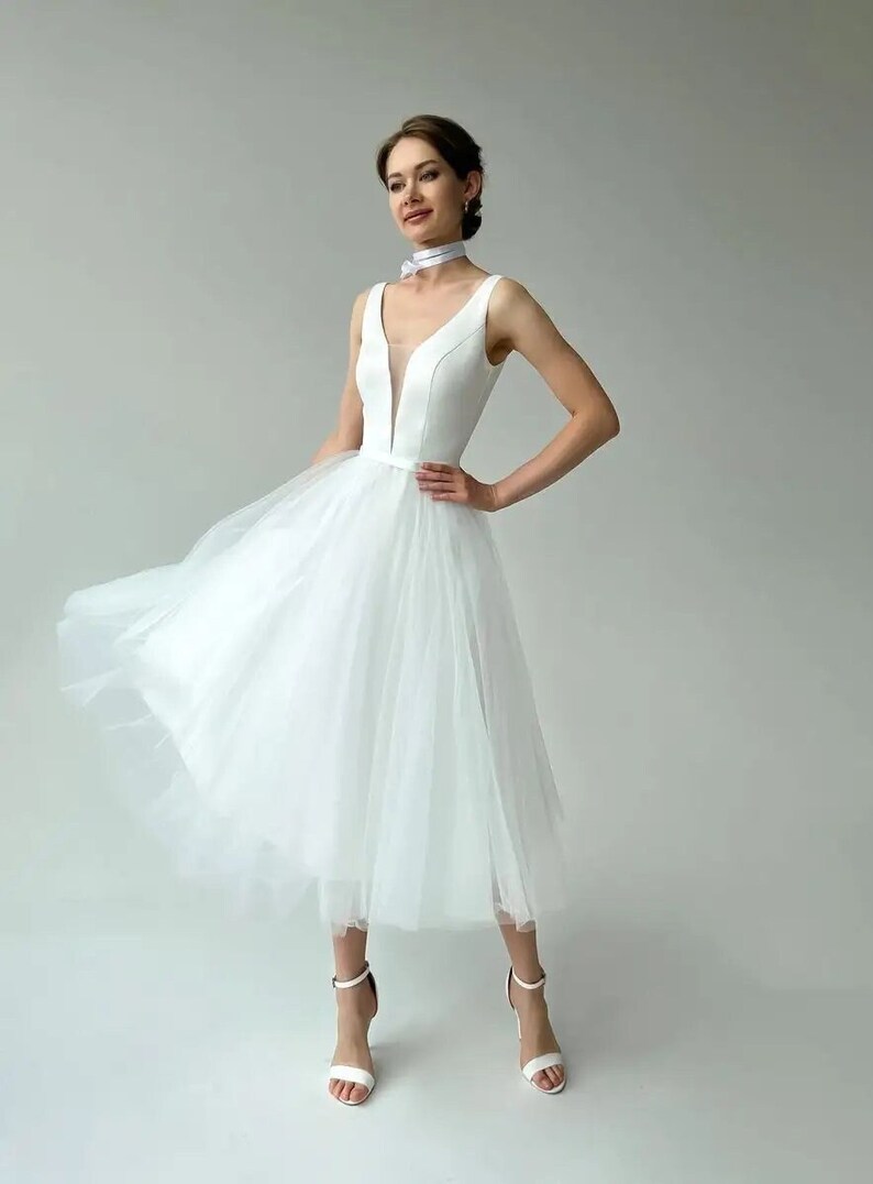 May include: A white knee-length dress with a V-neckline and a tulle skirt. The dress has a fitted bodice and a flowing skirt. The model is wearing white high-heeled sandals and a white choker. The dress is suitable for a wedding or special occasion.