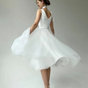 May include: A white knee-length dress with a V-neck and a layered tulle skirt. The dress is paired with white high-heeled sandals. The model is wearing a white ribbon around the neck. The dress is suitable for a wedding or special occasion.
