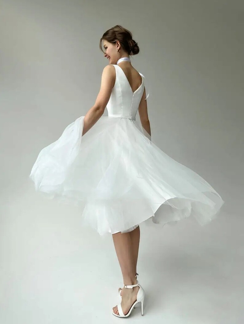 May include: A white knee-length dress with a V-neck and a layered tulle skirt. The dress is paired with white high-heeled sandals. The model is wearing a white ribbon around the neck. The dress is suitable for a wedding or special occasion.