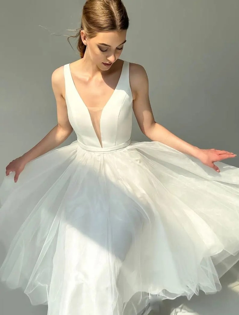 May include: A white wedding dress with a deep V-neckline and a flowing tulle skirt. The sleeveless bodice has a sheer panel at the chest. The dress is designed for a formal occasion.