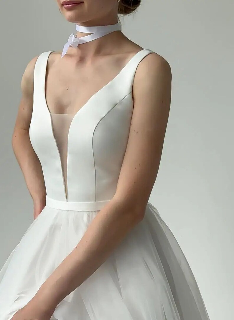 May include: A white wedding dress with a V-neckline and a sheer panel. The sleeveless bodice has a fitted silhouette, and the skirt is full and flowing. A white ribbon choker is worn around the neck.