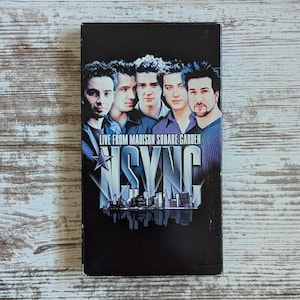 May include: A black VHS tape featuring the boy band *NSYNC. The cover art shows the band members' faces above the text "LIVE FROM MADISON SQUARE GARDEN" and the band's name. The background is a dark gradient.