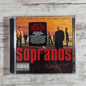 May include: A CD case for "The Sopranos" featuring the show's title in red, the phrase "Peppers & Eggs", and a Parental Advisory Explicit Lyrics warning. The cover art includes images of characters from the series.