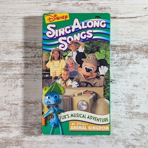 May include: A green VHS tape for Disney's Sing Along Songs: Flik's Musical Adventure at Disney's Animal Kingdom. The cover features Mickey Mouse in a safari hat, children, and the character Flik.