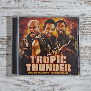 May include: A CD case featuring the movie "Tropic Thunder" soundtrack. The cover art shows three men in military attire, with the movie title in large, bold letters. The background is a fiery orange and red.