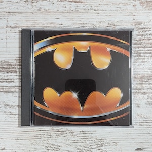May include: A square compact disc jewel case featuring a glossy black cover with a prominent, stylized gold and black bat emblem. The design includes metallic gold highlights and a subtle silver outline, with a small trademark symbol in the corner.
