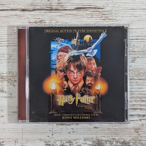 May include: A Harry Potter and the Sorcerer's Stone original motion picture soundtrack CD. The cover features Harry Potter, other characters, and a white owl. The title and composer's name are also visible.