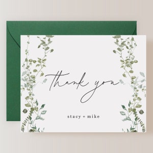 May include: White thank you card with green watercolor foliage border. The text "Thank you" is written in black cursive script. Below the text is "Stacy + Mike" in black text.