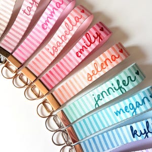 Op de afbeelding: Een verzameling gestreepte pols keychains in verschillende kleuren, elk met een naam geborduurd in een bijpassende kleur. Keychains bevatten namen als Mia, Taylor, Emma, Isabella, Emily, Selena, Jennifer, Megan, Kylie en Olivia.