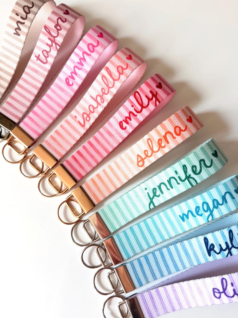 Op de afbeelding: Een verzameling gestreepte pols keychains in verschillende kleuren, elk met een naam geborduurd in een bijpassende kleur. Keychains bevatten namen als Mia, Taylor, Emma, Isabella, Emily, Selena, Jennifer, Megan, Kylie en Olivia.
