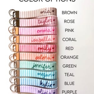 Op de afbeelding: Een selectie gestreepte pols-sleutelhangers in verschillende kleuren, elk met een naam en een hart. De kleuropties zijn bruin, roze, roze, koraal, rood, oranje, groen, blauwgroen, blauw en paars. De tekst "COLOR OPTIONS" staat bovenaan.