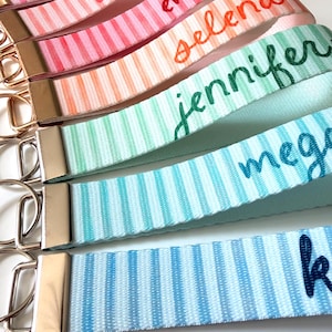 Op de afbeelding: Close-up van verschillende gepersonaliseerde sleutelhangers met streepjespatronen in verschillende pastelkleuren. Elke sleutelhanger heeft een andere naam in een cursief lettertype, waaronder "Emily", "Selena" en "Jennifer". De sleutelhangers hebben goudkleurige hardware.