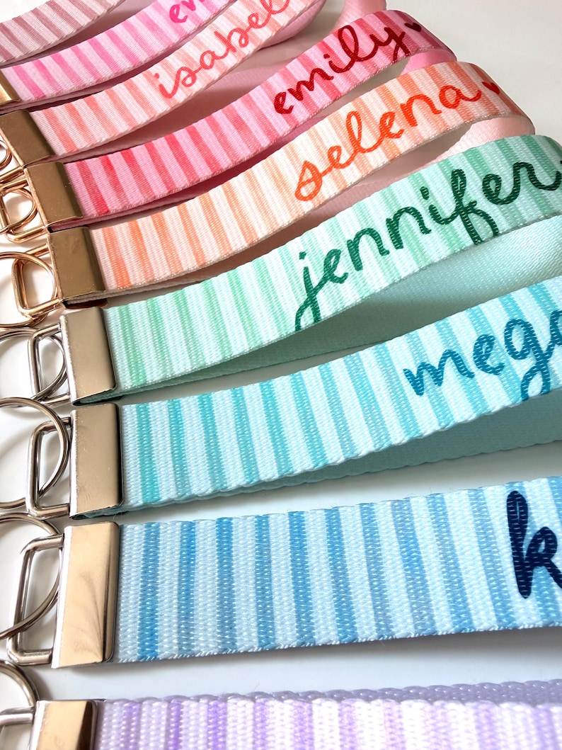 Op de afbeelding: Close-up van verschillende gepersonaliseerde sleutelhangers met streepjespatronen in verschillende pastelkleuren. Elke sleutelhanger heeft een andere naam in een cursief lettertype, waaronder "Emily", "Selena" en "Jennifer". De sleutelhangers hebben goudkleurige hardware.