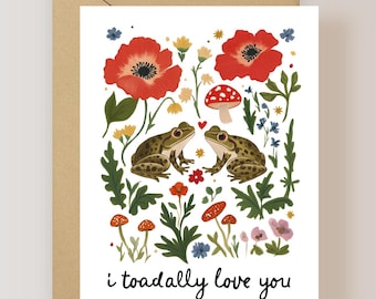 May include: A white greeting card featuring two green spotted frogs facing each other, surrounded by red poppies, small yellow flowers, and red-capped mushrooms. A small red heart sits between the frogs. The text reads: i toadally love you.