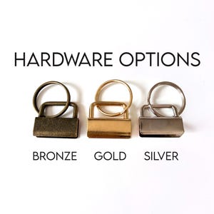 Op de afbeelding: Afbeelding van drie sleutelhanger hardware opties: brons, goud en zilver. Elke optie bevat een rechthoekige sluiting en een ronde sleutelring. De tekst "HARDWARE OPTIONS" staat boven de hardware, met de kleurnamen eronder.