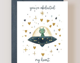 May include: A white greeting card featuring a dark blue UFO with a green alien inside. The text reads you've abducted my heart. The design includes gold hearts, stars, and sparkles scattered around the central flying saucer illustration.