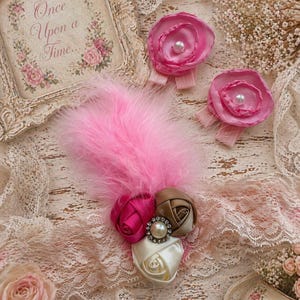 May include: A collection of pink and cream-colored fabric flower hair clips and embellishments. One clip features a cluster of satin roses with a pearl and rhinestone center. A pink feather and a framed print with the text "Once Upon a Time..." are also present.
