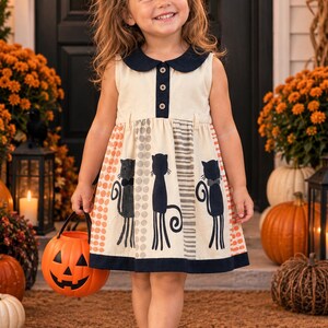 May include: A sleeveless cream-colored dress featuring a dark navy blue Peter Pan collar, a button-down bodice, and a skirt decorated with vertical stripes of orange dots, gray circles, and three black cat silhouettes with long curled tails.