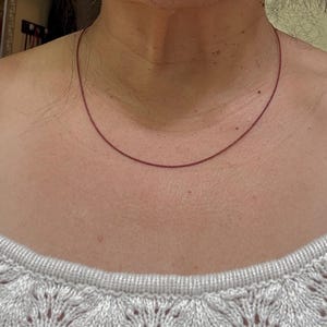 Puede incluir: Un delicado y fino collar rojo sobre el cuello de una persona. El collar es de diseño simple y minimalista, probablemente hecho de un cordón fino. La persona lleva un suéter blanco de punto abierto. El collar mide unos 40 cm.