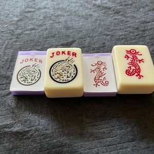 May include: Four ivory-colored Mahjong tiles with purple edges. Two tiles read "JOKER" with dragon graphics, one in white and one in red. The other two tiles feature red dragon designs. The tiles are rectangular and have rounded edges.
